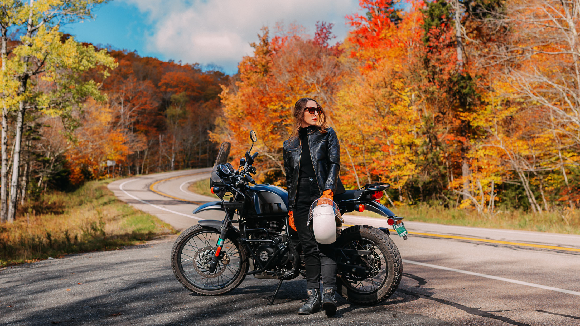 A rider in a black leather jacket stands beside a Royal Enfield Himalayan at Middlebury Gap, holding a white helmet as the road curves through bright autumn foliage.