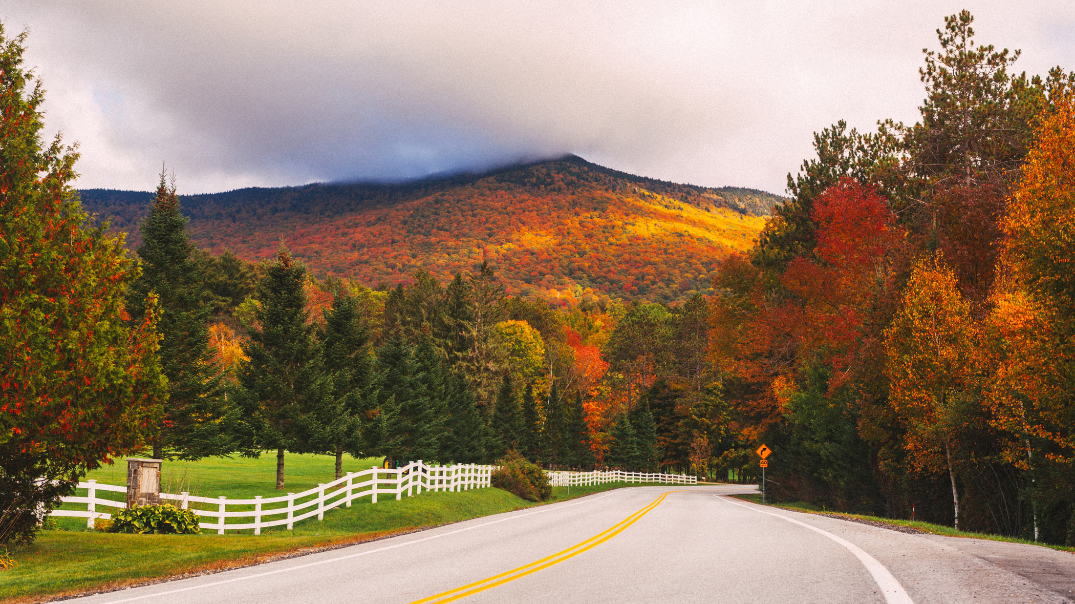 A two-lane road curves past a white fence toward a mountain covered in autumn foliage, with low clouds along the ridge.