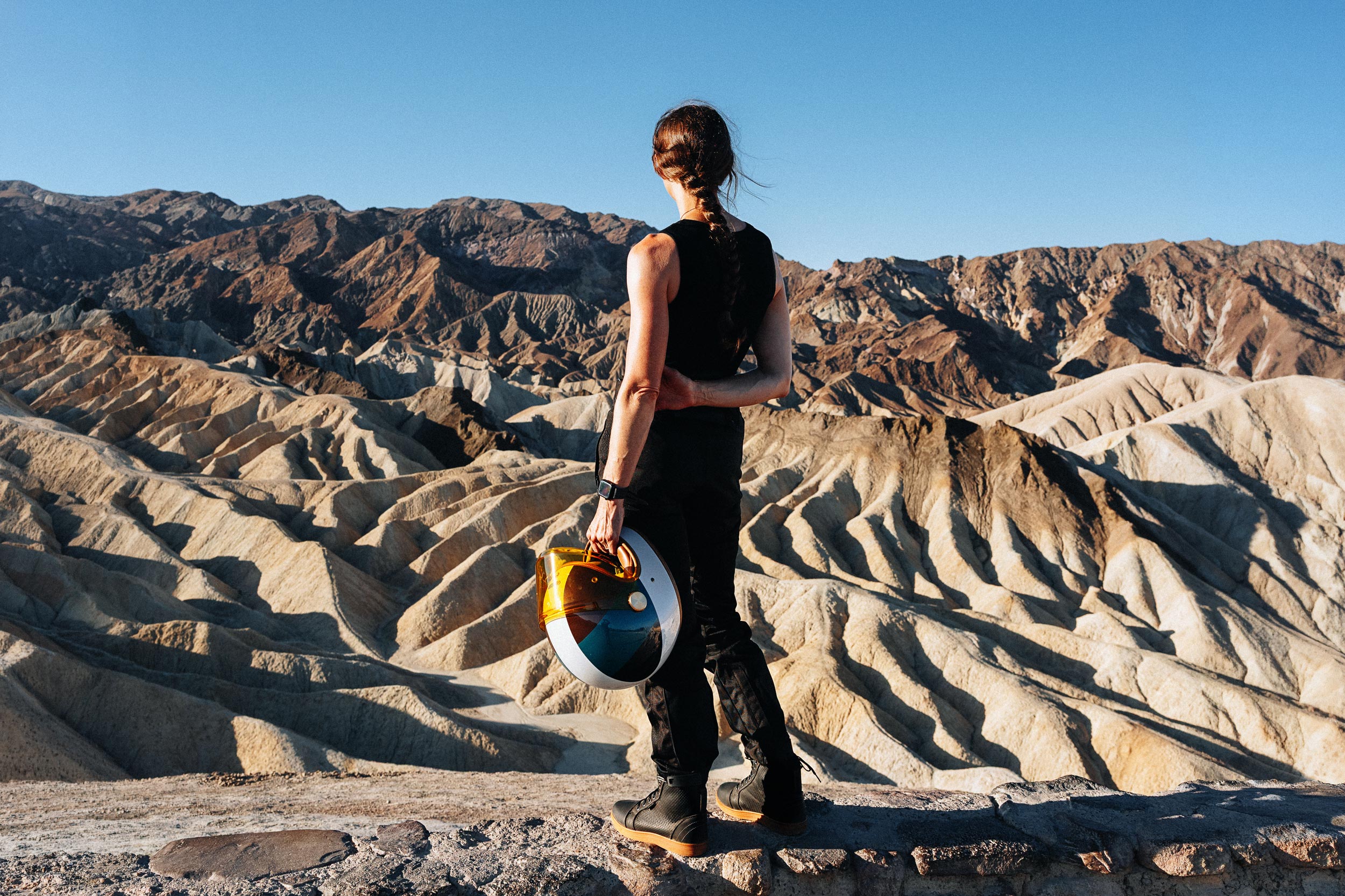 Woman wearing black Indie Ridge Crossfield ventilated motorcycle boots and holding a colorful helmet at Zabriskie Point in Death Valley.