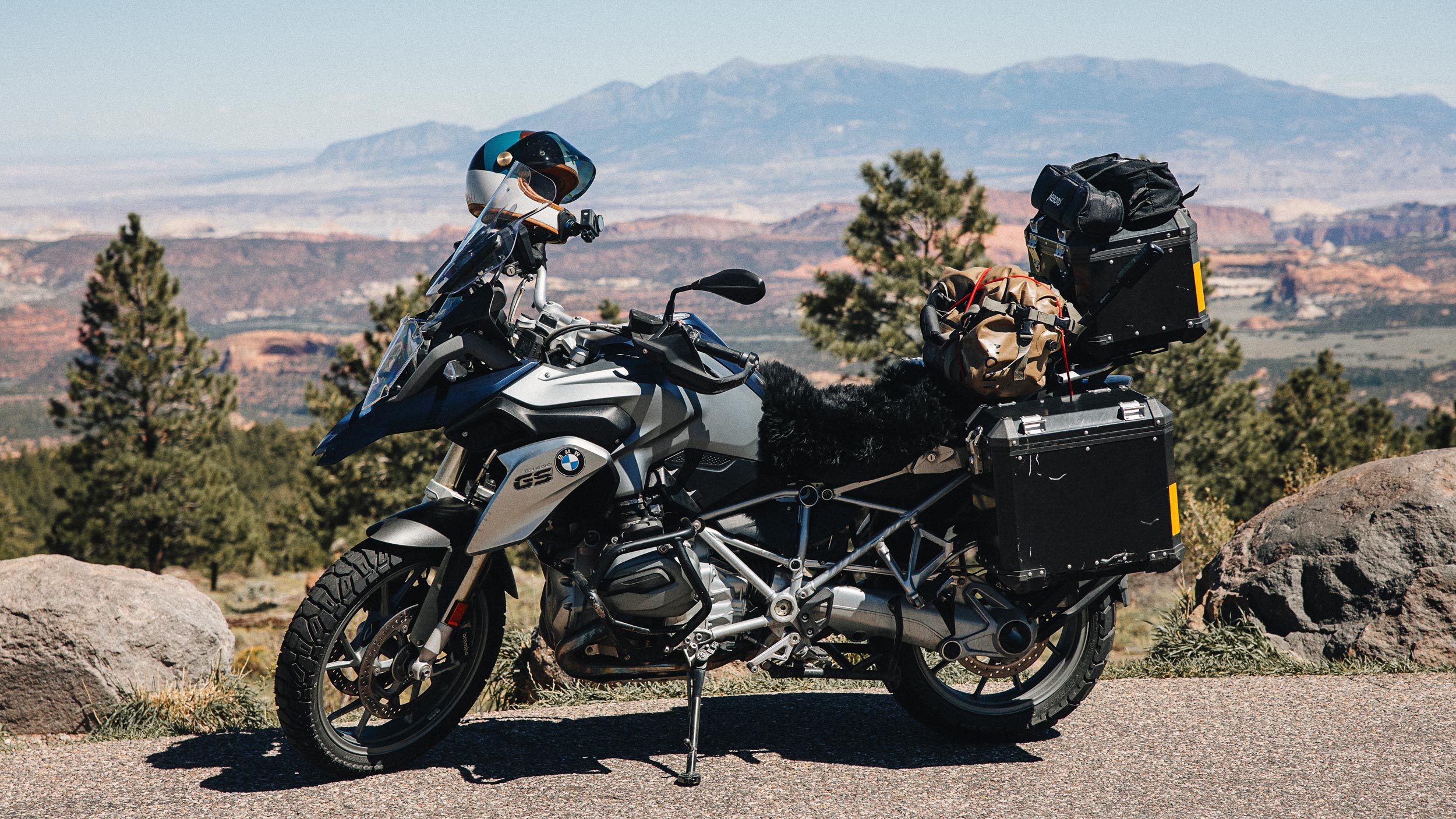 Fully loaded BMW GS motorcycle with hard panniers, camping bags, a helmet, and a black sheepskin parked at a scenic mountain overlook.