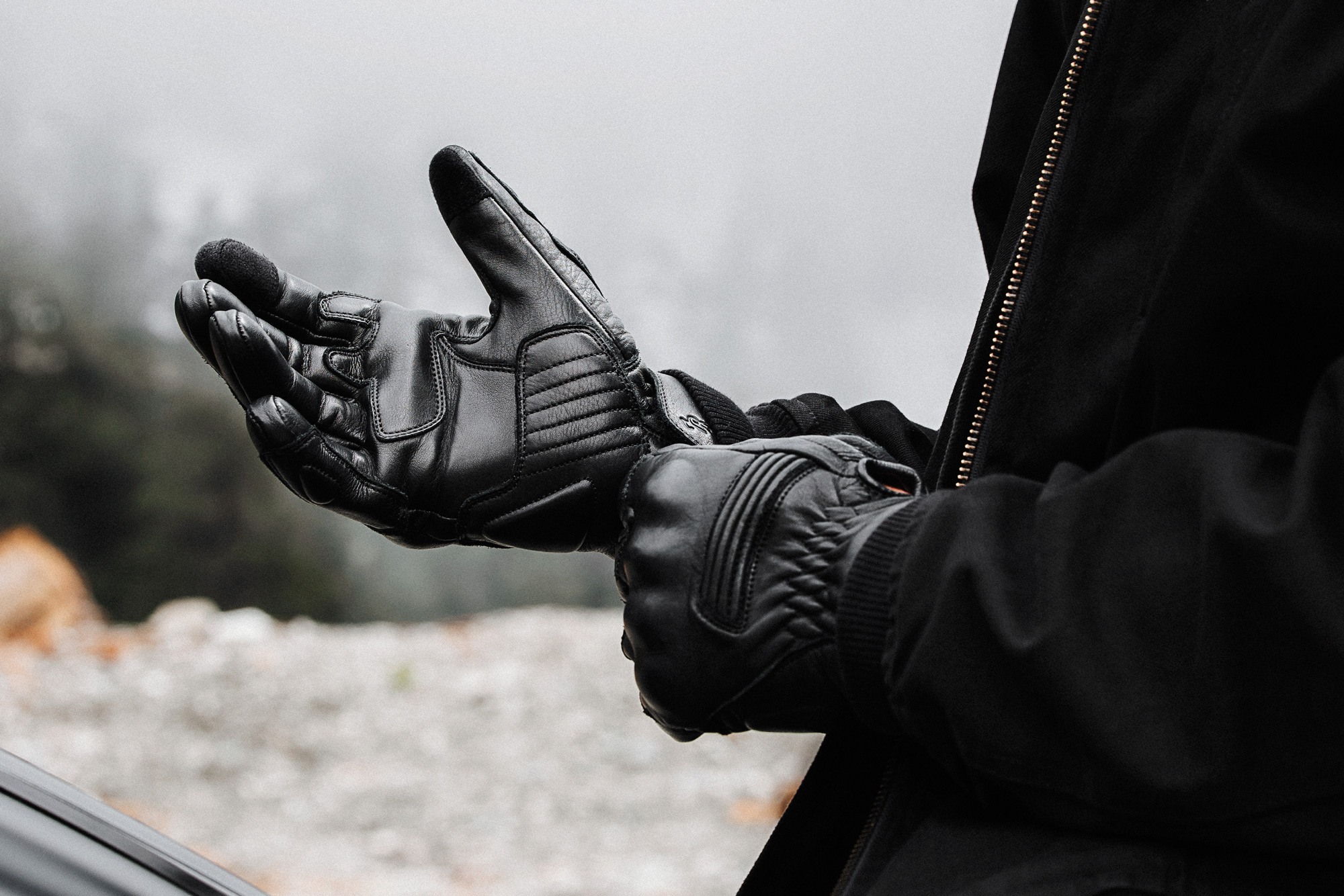A motorcyclist pulls on black leather motorcycle gloves before a ride on a cool, foggy morning, highlighting the importance of quality riding gear and proper preparation.