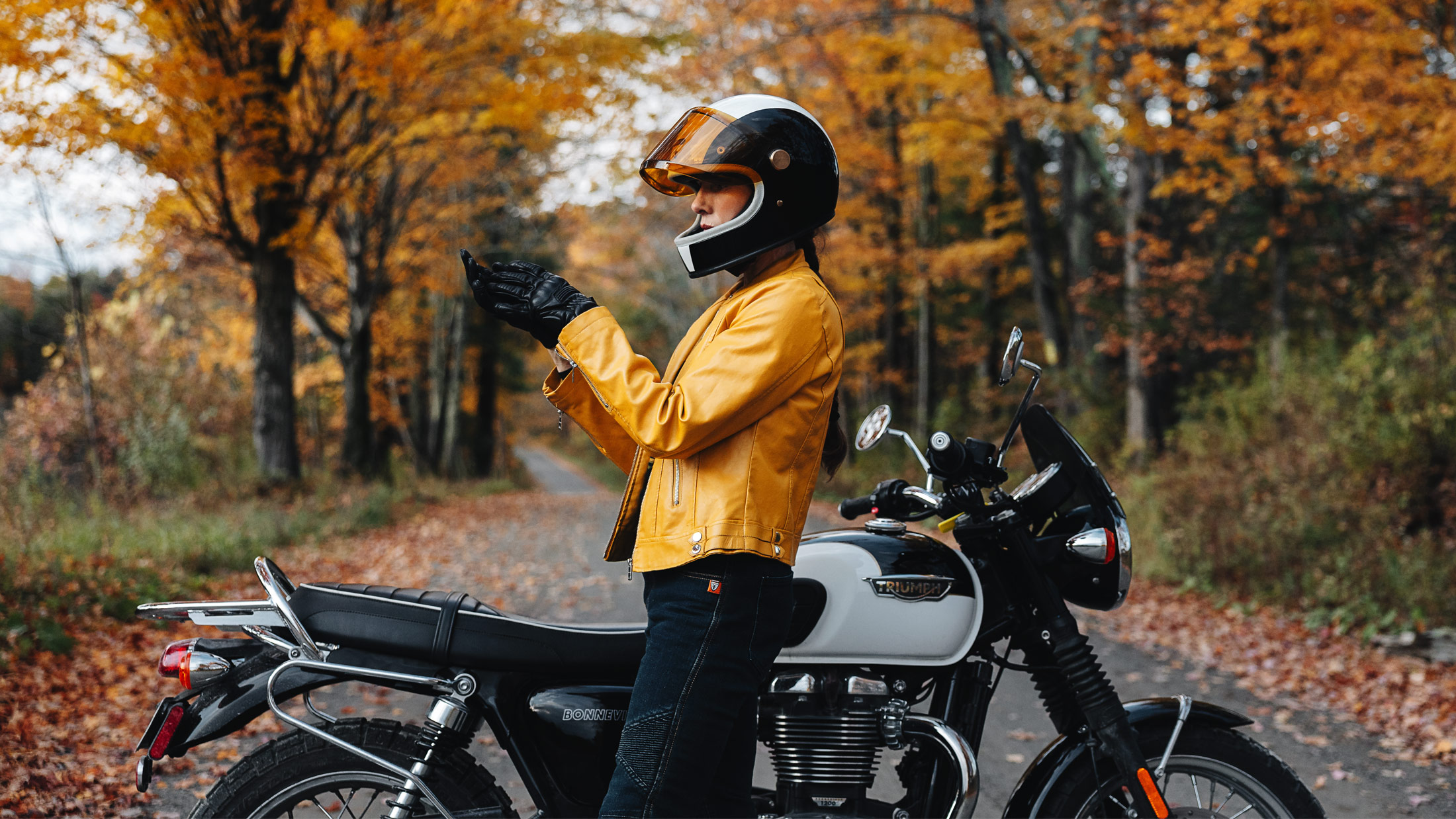 Caption: Pulling on my gloves before heading out for a fall ride. Alt text: A rider in a black helmet and mustard jacket adjusts a black leather glove beside a white Triumph Bonneville on an autumn road.