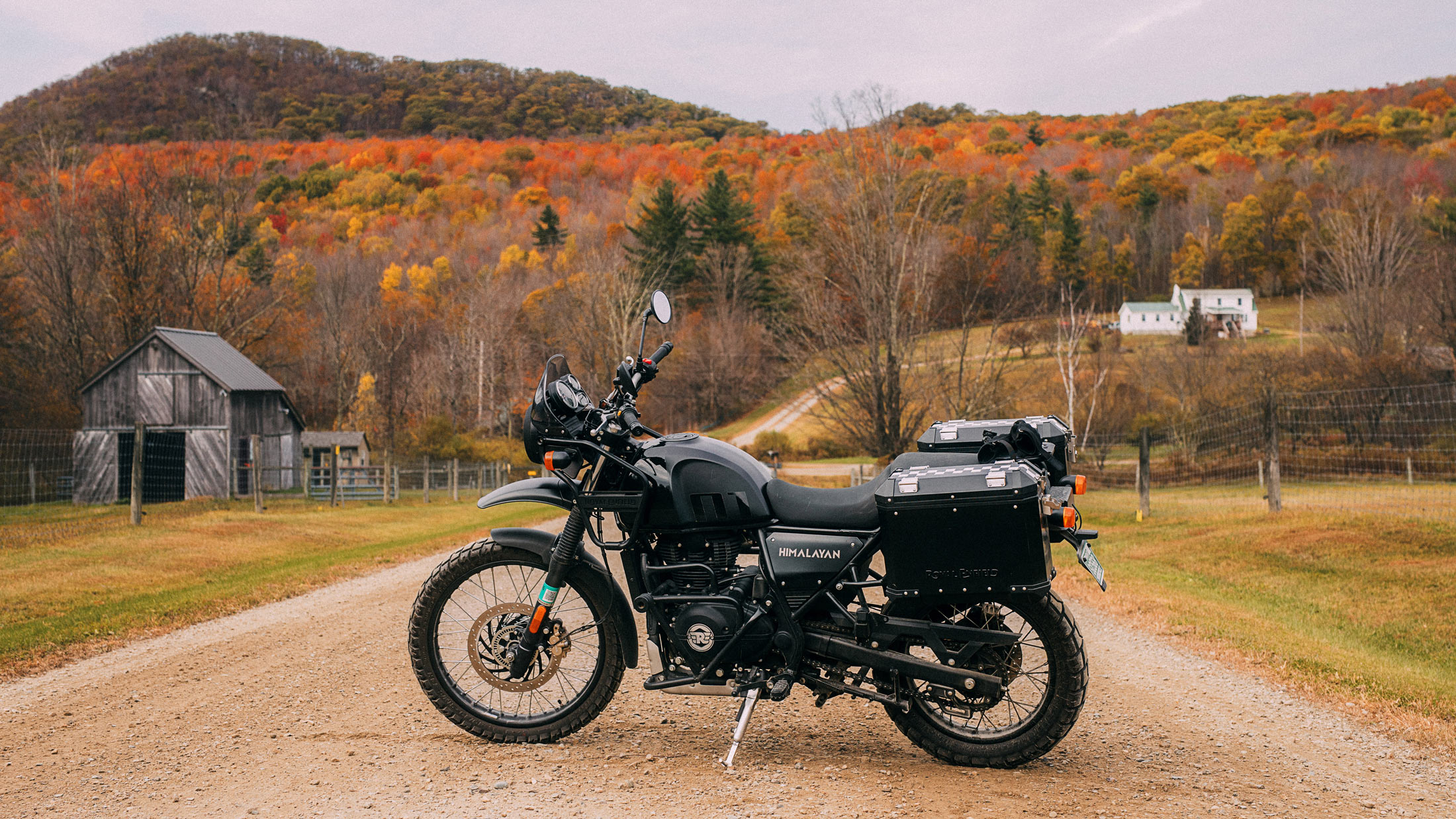 **Caption:** *A gravel-road detour on a fall ride in Vermont.*  **Alt text:** A black Royal Enfield Himalayan with luggage parked on a gravel road, with autumn foliage and farm buildings in the background.