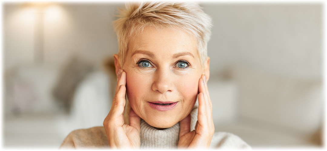 A smiling older woman with short blonde hair touches her face, highlighting her vibrant skin. Lily & Loaf showcases effective skincare for all ages.