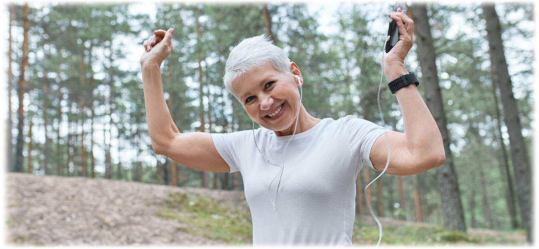 Active older woman with short gray hair smiling and raising her arms in triumph while holding a smartphone. She is wearing earphones and a fitness tracker in a forest setting, expressing joy and vitality.
