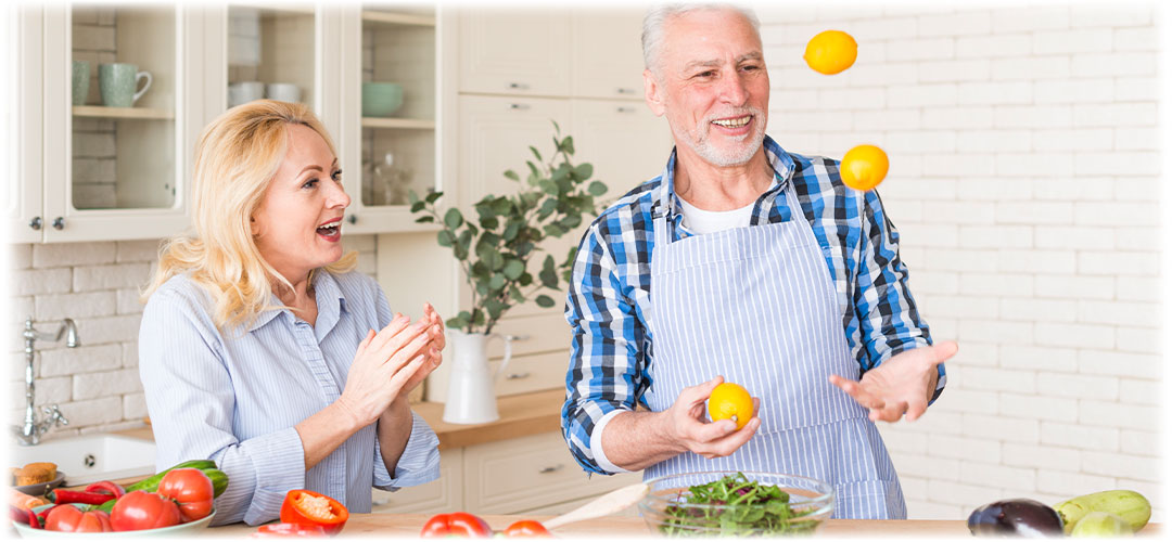 Smiling older couple in a bright kitchen. The man wears an apron and juggles oranges while the woman applauds joyfully. Fresh vegetables are laid out on the counter, creating a lively and playful cooking atmosphere.