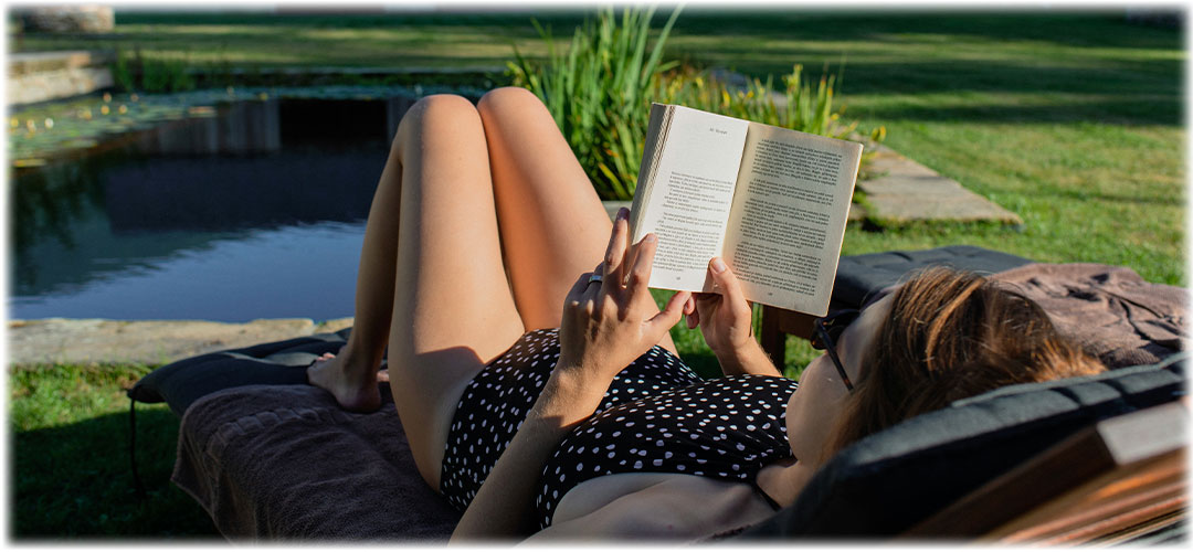 Woman in a black and white polka dot swimsuit relaxing on a lounge chair by a pond, reading a book under the warm sunlight. The scene evokes a sense of tranquility and leisure