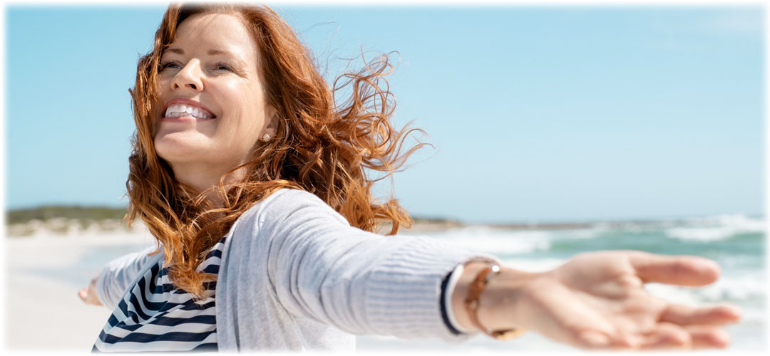 Smiling woman with red hair standing on a beach with her arms outstretched, enjoying the fresh sea breeze. She radiates happiness and freedom.