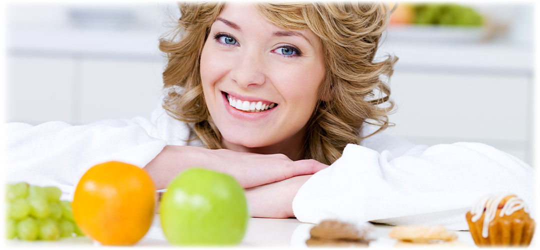 Smiling woman leaning on a white table surrounded by fresh fruits and healthy snacks. She exudes positivity and well-being in a bright kitchen setting.
