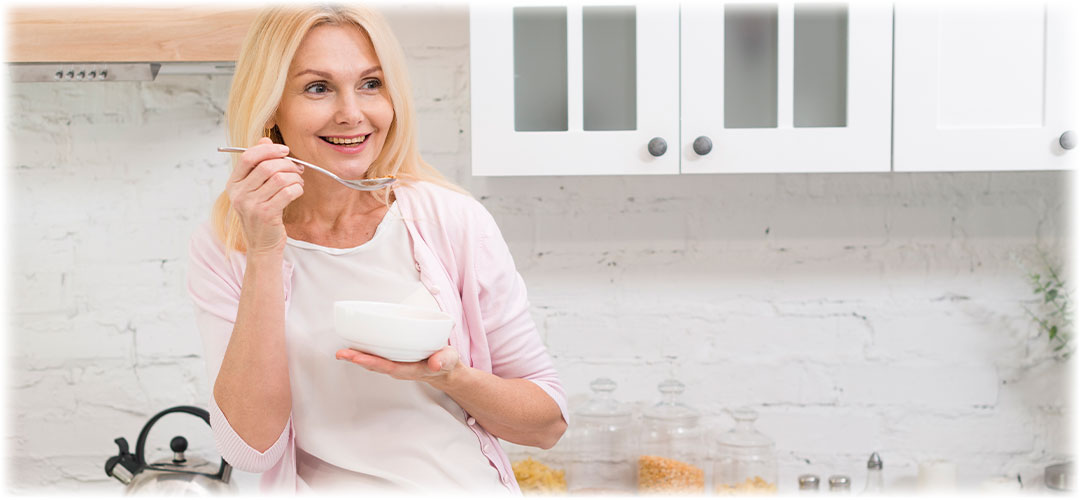 Smiling middle-aged woman eating from a white bowl with a spoon in a bright kitchen.