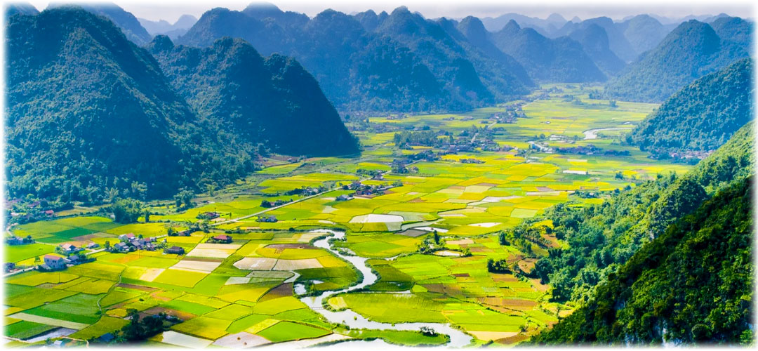 Aerial view of a lush, vibrant valley surrounded by towering mountains. The valley is dotted with colorful patchwork fields, small villages, and a winding river.