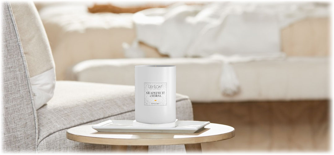 A Lily & Loaf Grapefruit and Mimosa scented candle sits on a tray atop a round table in a cozy, light-filled living room. Lily & Loaf promotes relaxation and ambiance.