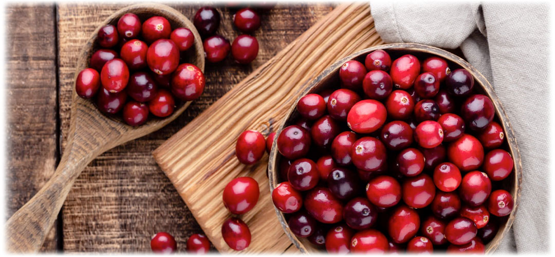 Top view of a wooden bowl and spoon filled with fresh cranberries on a rustic wooden surface. The rich red berries symbolize natural health benefits and vibrant flavor.