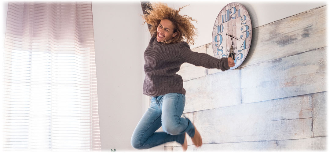 Joyful woman with curly hair jumping on a bed with a big smile. She wears a cozy sweater and jeans, expressing excitement and carefree happiness in a bright, well-lit bedroom.