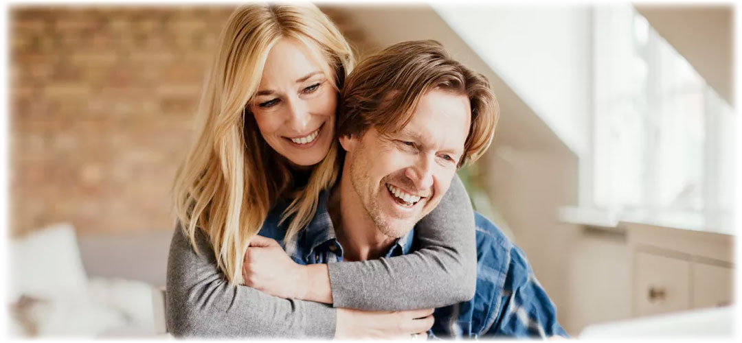 Happy couple embracing and smiling warmly in a cozy home setting. The woman wraps her arms around the man, both radiating love and joy.