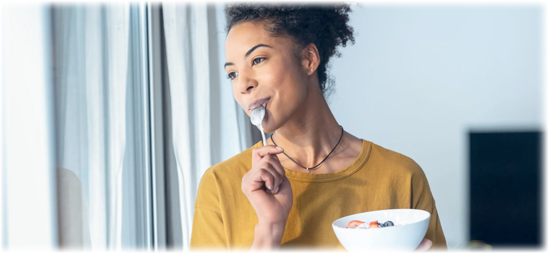 A young woman with curly hair enjoys a bowl of yogurt with fresh fruit, smiling thoughtfully by a window. Lily & Loaf promotes healthy and mindful eating.