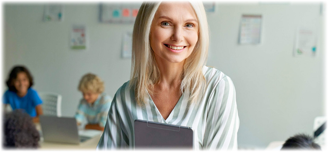 A smiling blonde woman holding a clipboard stands in a classroom with children working on laptops in the background. Lily & Loaf supports education and learning.