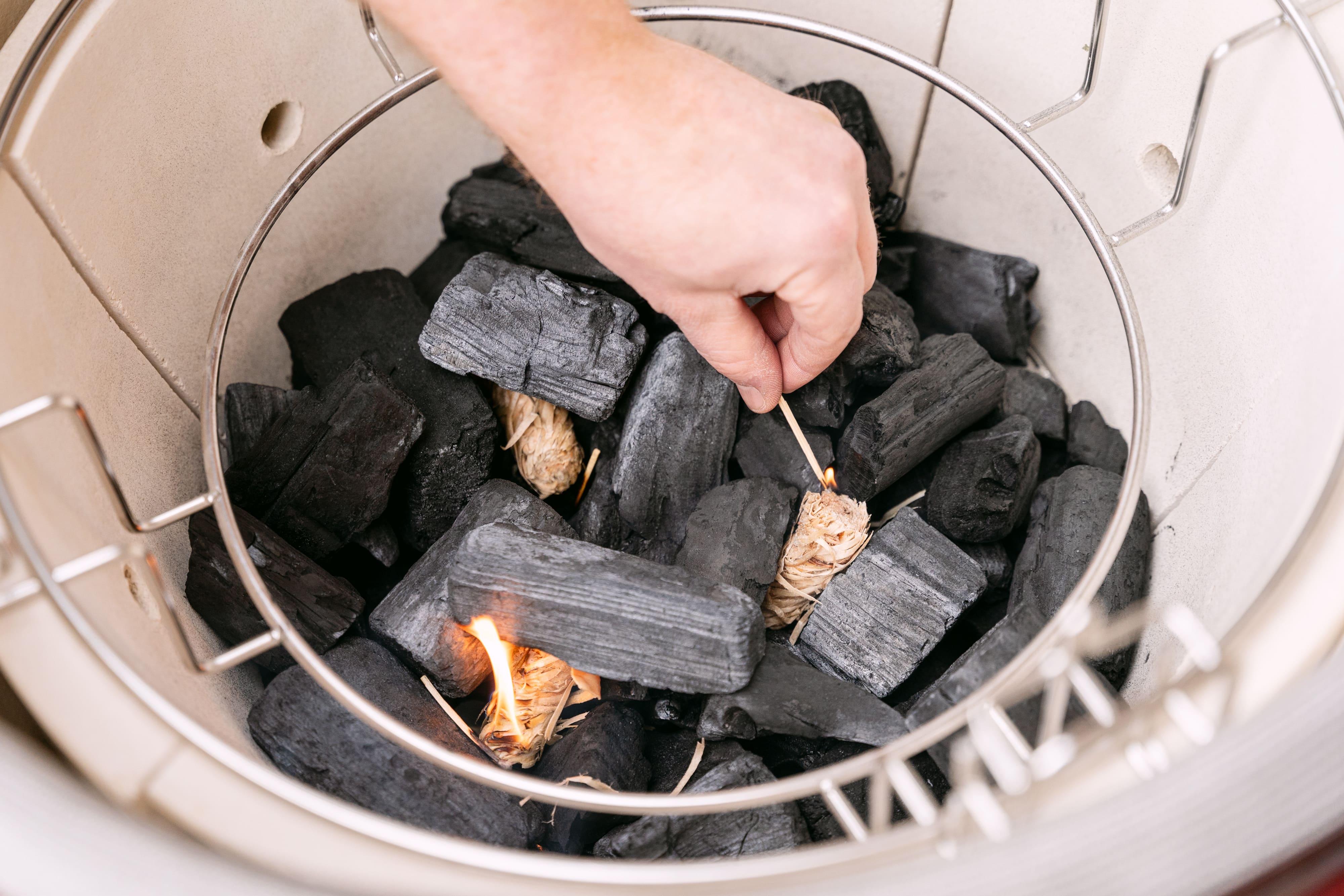Une personne allume du charbon de bois dans un Kamado en utilisant un briquet, montrant l'intérieur rempli de charbon et de morceaux de bois enflammés pour préparer une cuisson au barbecue.
