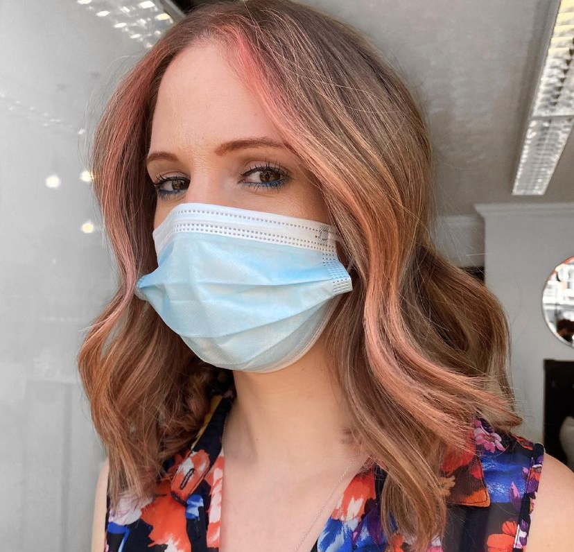 woman wearing a face mask looking head on into camera with brown hair and pink pieces at the front